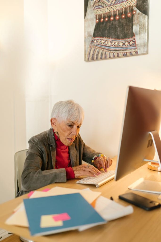 an-elderly-woman-in-gray-blazer-typing-on-computer-keyboard-8124377 An elderly woman in a gray blazer typing on a computer keyboard at her desk in a modern office setting.