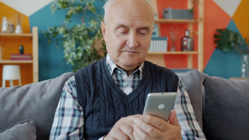 elderly-man-using-a-smartphone-on-a-couch-4m7hln1r6ss Elderly man is using smartphone touching screen sitting on couch in house alone enjoying modern device. Communication, gadgets and old people concept.